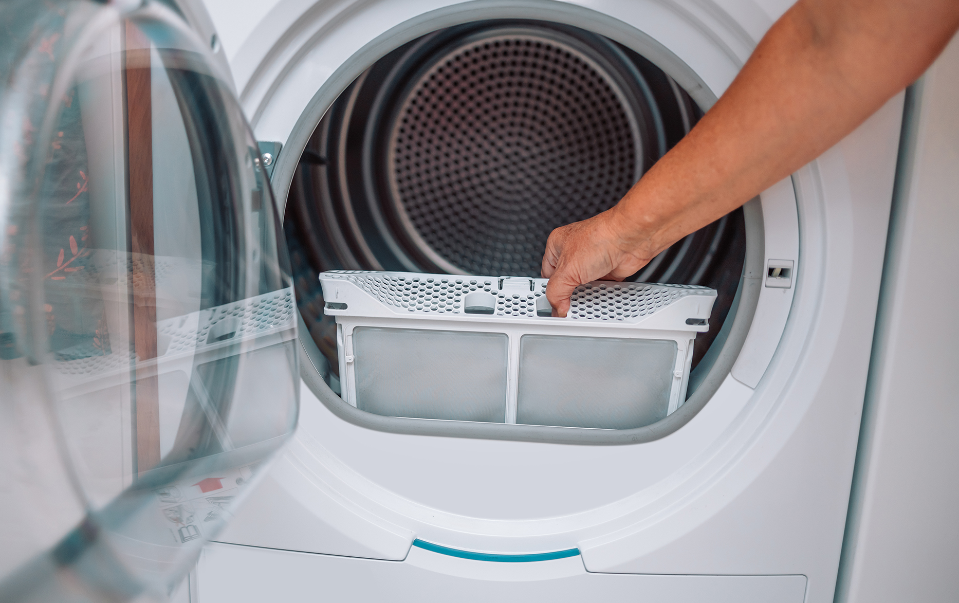 A real photo of a home laundry room with a front-load dryer door open and a hand pulling out the lint screen, natural indoor lighting