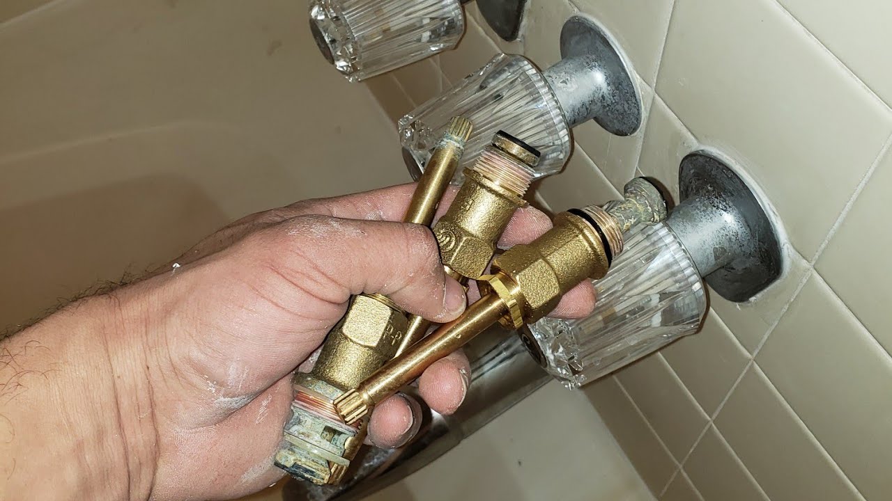 A real photo of a homeowner removing a brass diverter stem from a three-handle tub valve with a deep socket, with the trim plate visible