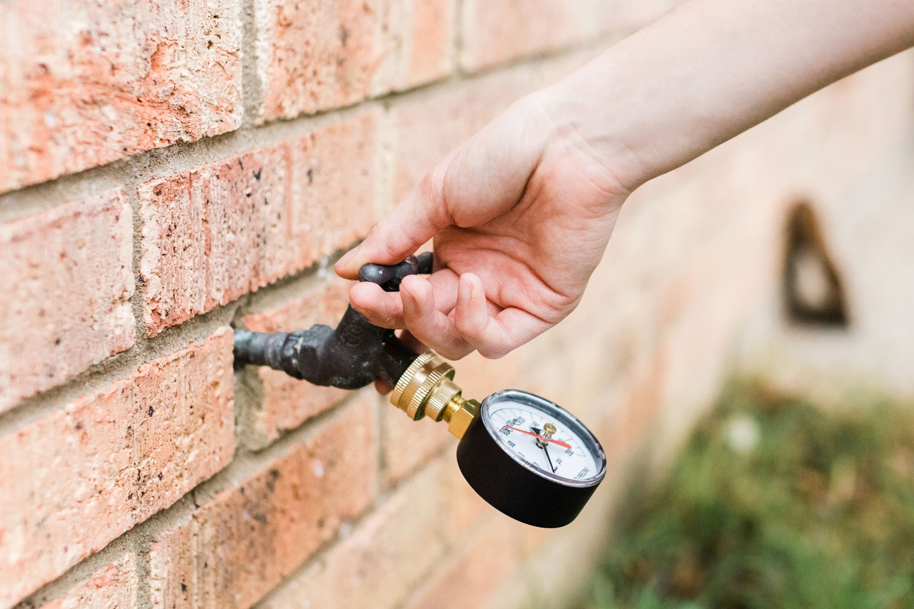 A real photo of a homeowner’s hand threading a round water pressure gauge onto an outdoor hose bib on a brick exterior wall in daylight, close-up plumbing test scene