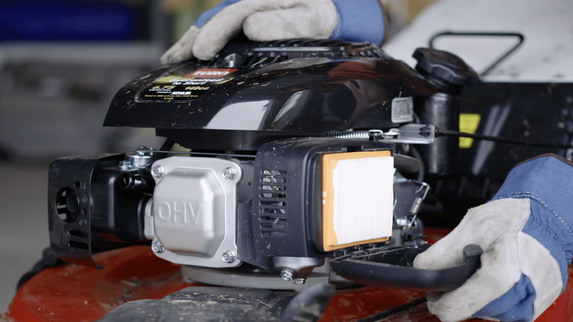 A real photo of a lawn mower with the air filter cover removed, showing the filter element in the housing on a workbench