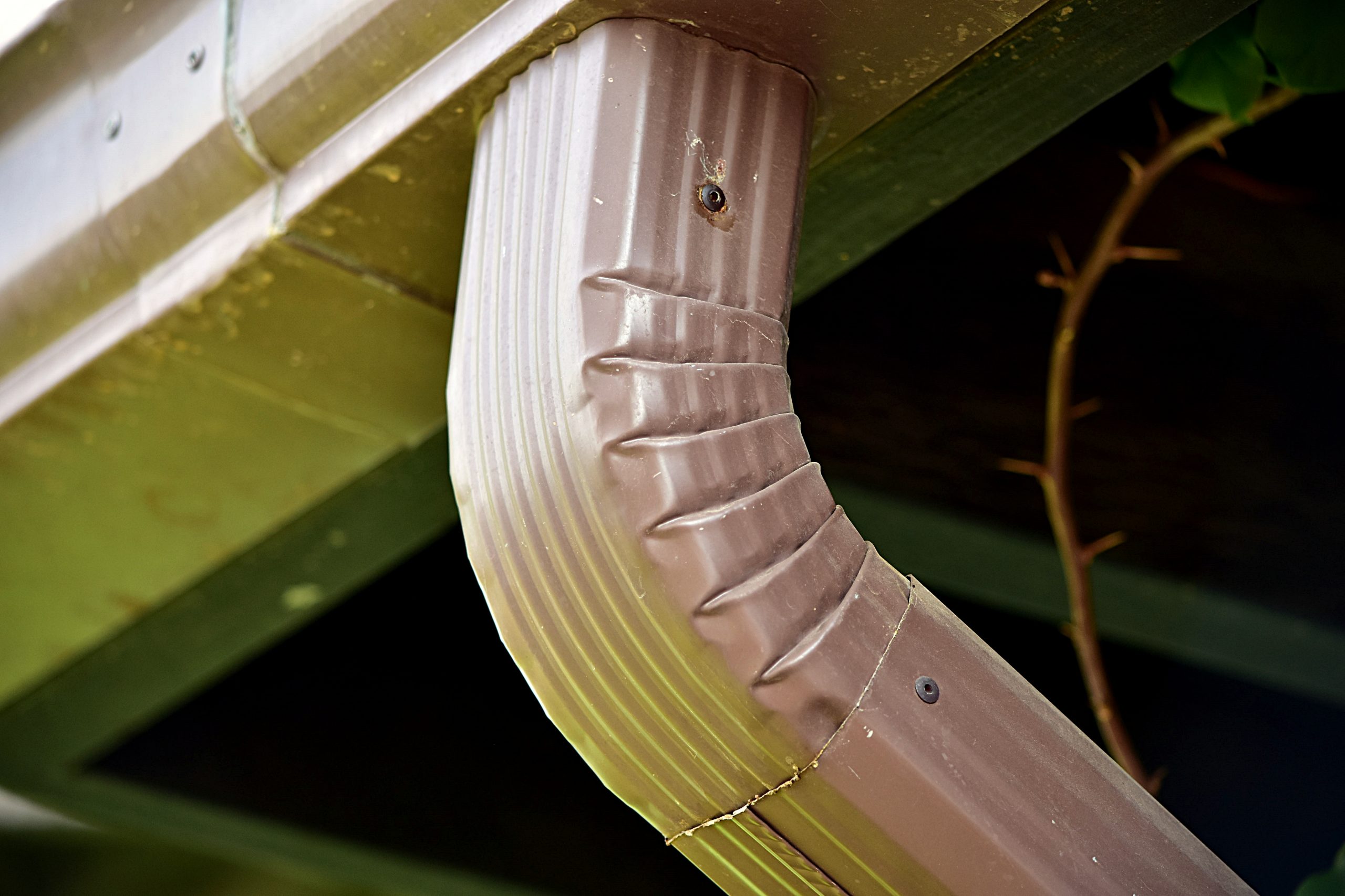 A real photo of a metal downspout elbow connection on a house exterior secured with small sheet metal screws in daylight
