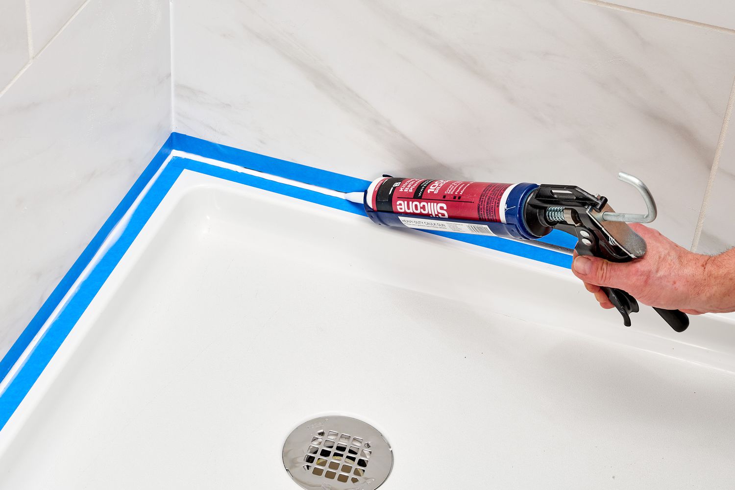 A real photo of a person applying clear 100% silicone caulk with a caulk gun in the vertical corner seam of a tiled shower