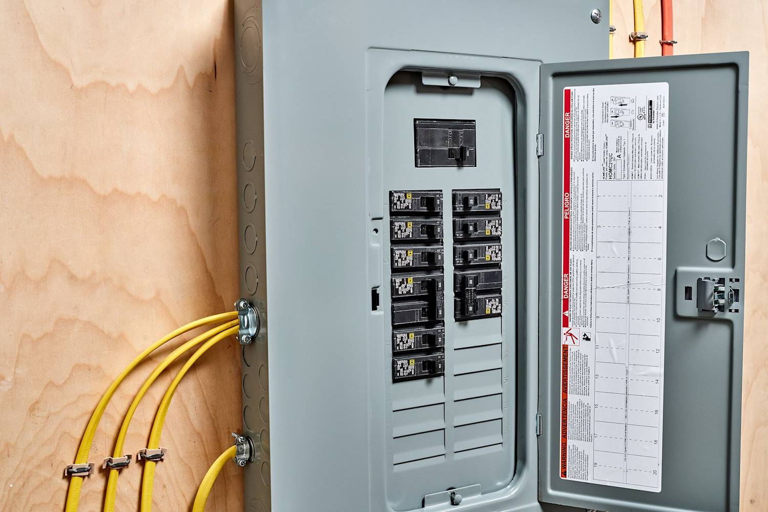 A real photo of a residential electrical breaker panel with the door open, showing rows of breakers in a utility area with soft indoor lighting