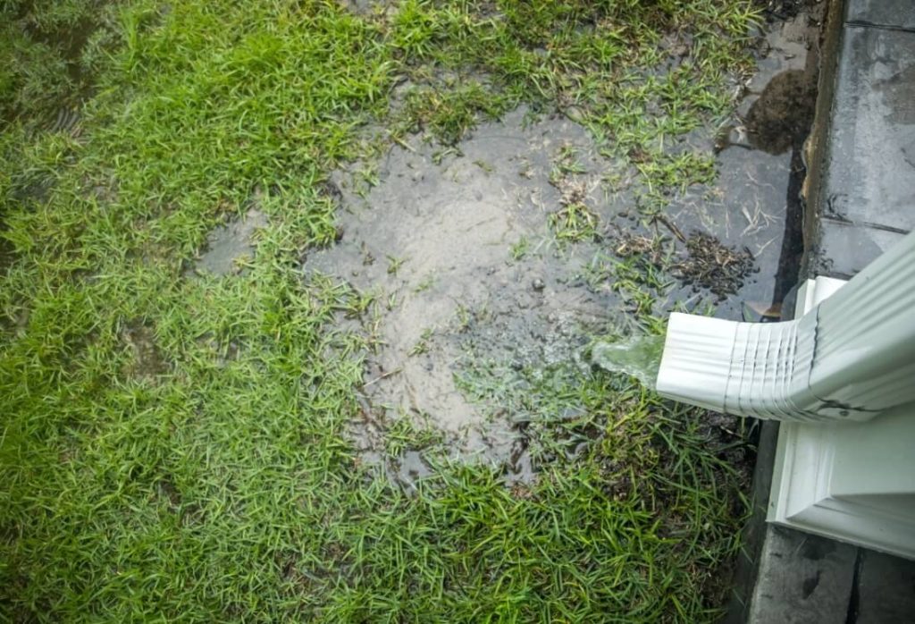 A real photo of a roof downspout releasing water directly onto soil next to a house foundation during wet weather, with puddling visible