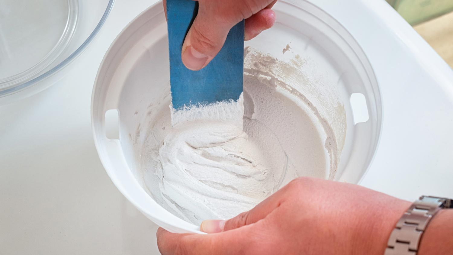 A real photo of a small bucket with grout being mixed to a thick paste using a margin trowel, with shower tile in the background