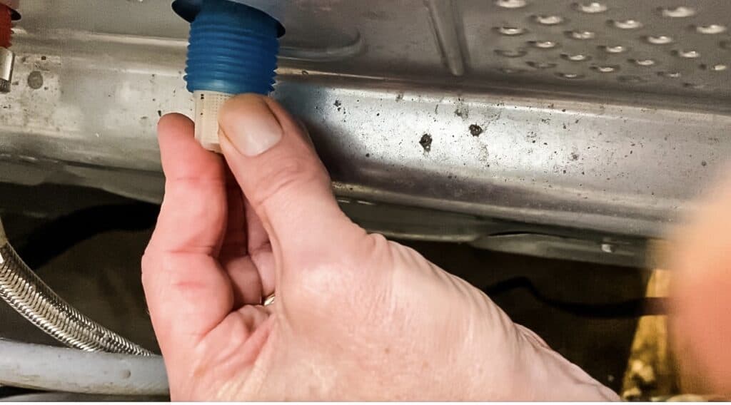 A real photo of a washing machine water inlet connection with the hose removed, showing a person cleaning the small mesh screen with a soft brush