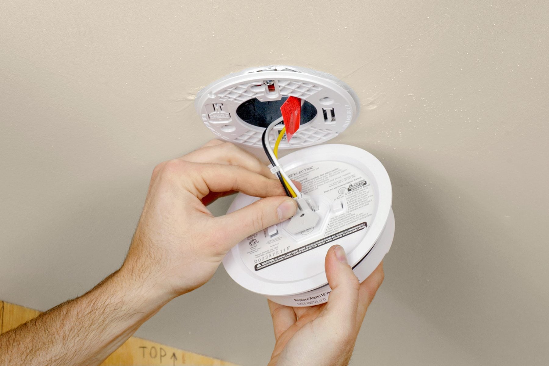 A real photo of a white hardwired smoke detector mounted on a ceiling with the battery door open and a 9-volt battery visible