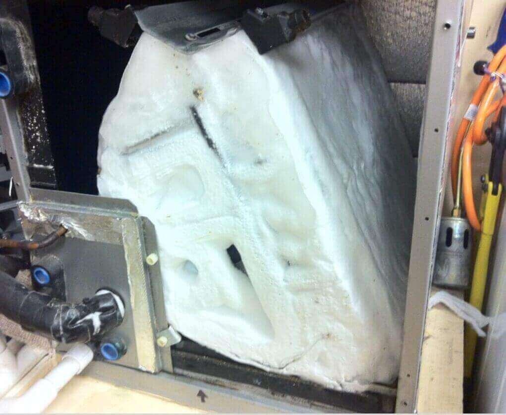 A real photo of an indoor evaporator coil inside an air handler covered in thick white ice and frost, with the access panel removed in a home utility area