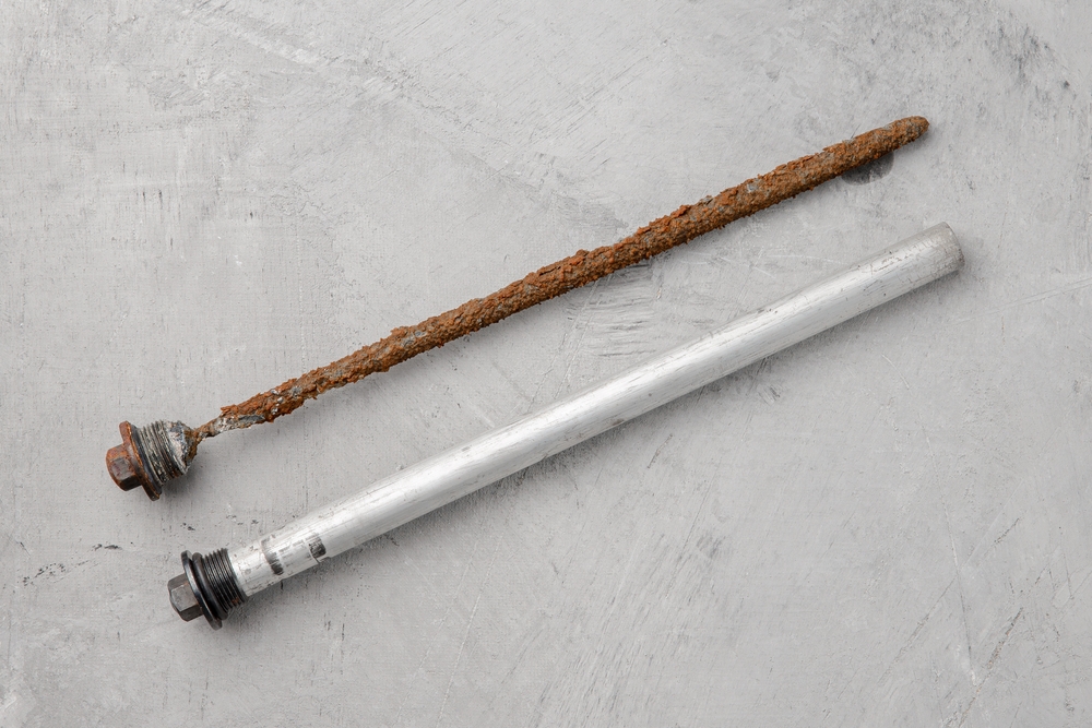 A real photo of different water heater anode rods laid on a workbench, including a straight magnesium rod and a segmented flexible rod, in a garage workshop
