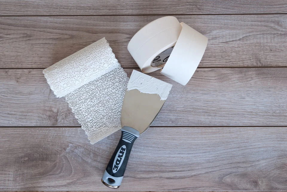 A real photo of drywall repair tools on a drop cloth, including joint compound, putty knives, sanding sponge, and drywall screws in a small container