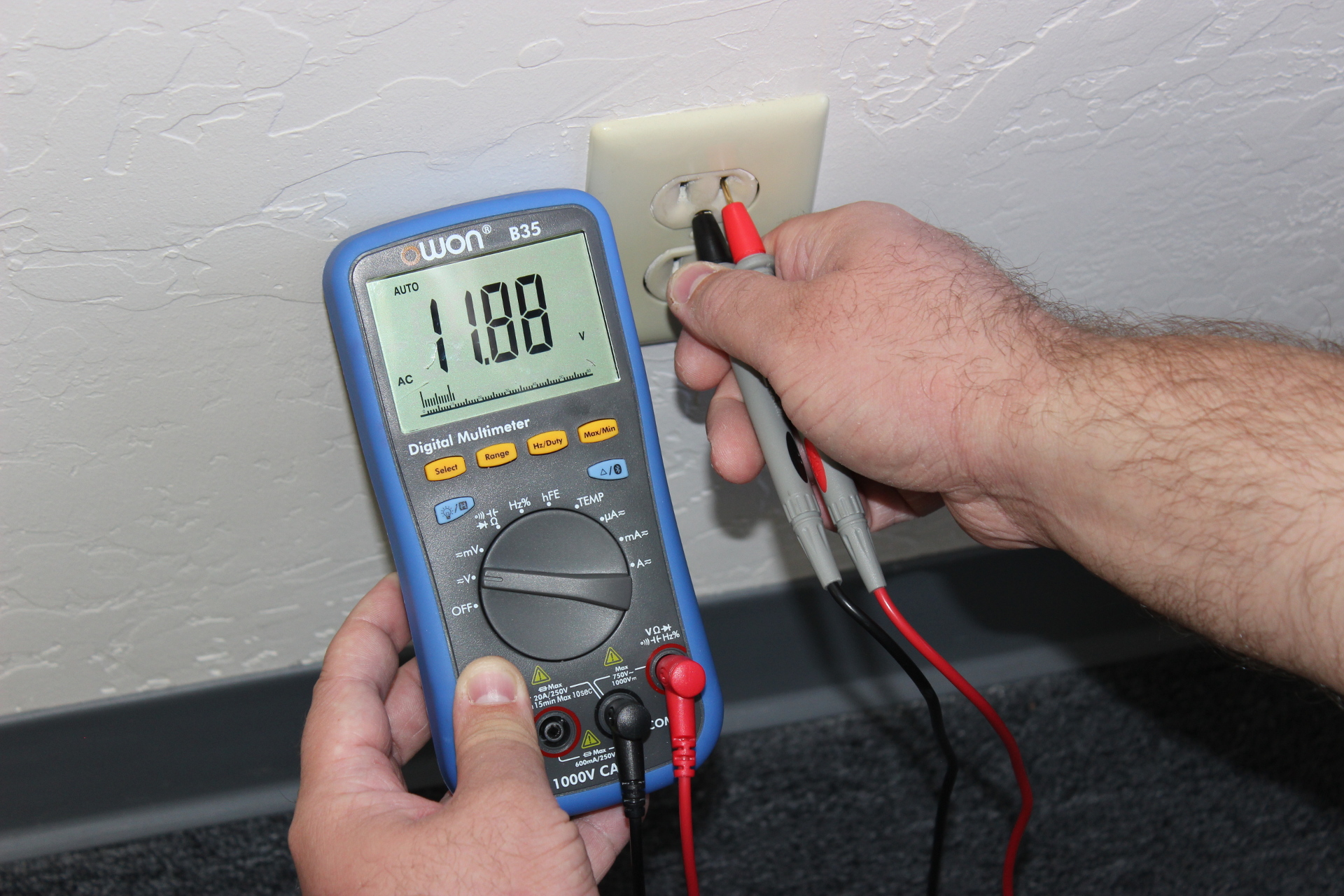 A real photo of hands holding multimeter probes near an electrical outlet on a wall, with the multimeter screen visible and the probes positioned carefully, indoor home lighting