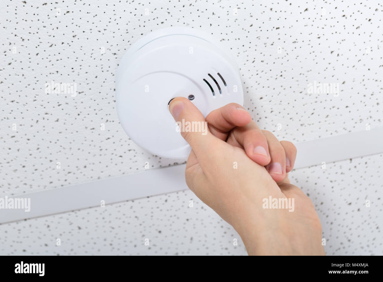 A real photo of hands twisting a round smoke detector off its ceiling mounting bracket in a residential hallway