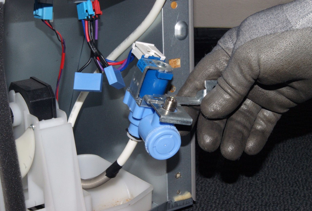 A real photo of the lower back of a refrigerator pulled slightly away from the wall, showing the water inlet valve assembly with connected water line and electrical plugs, indoor lighting