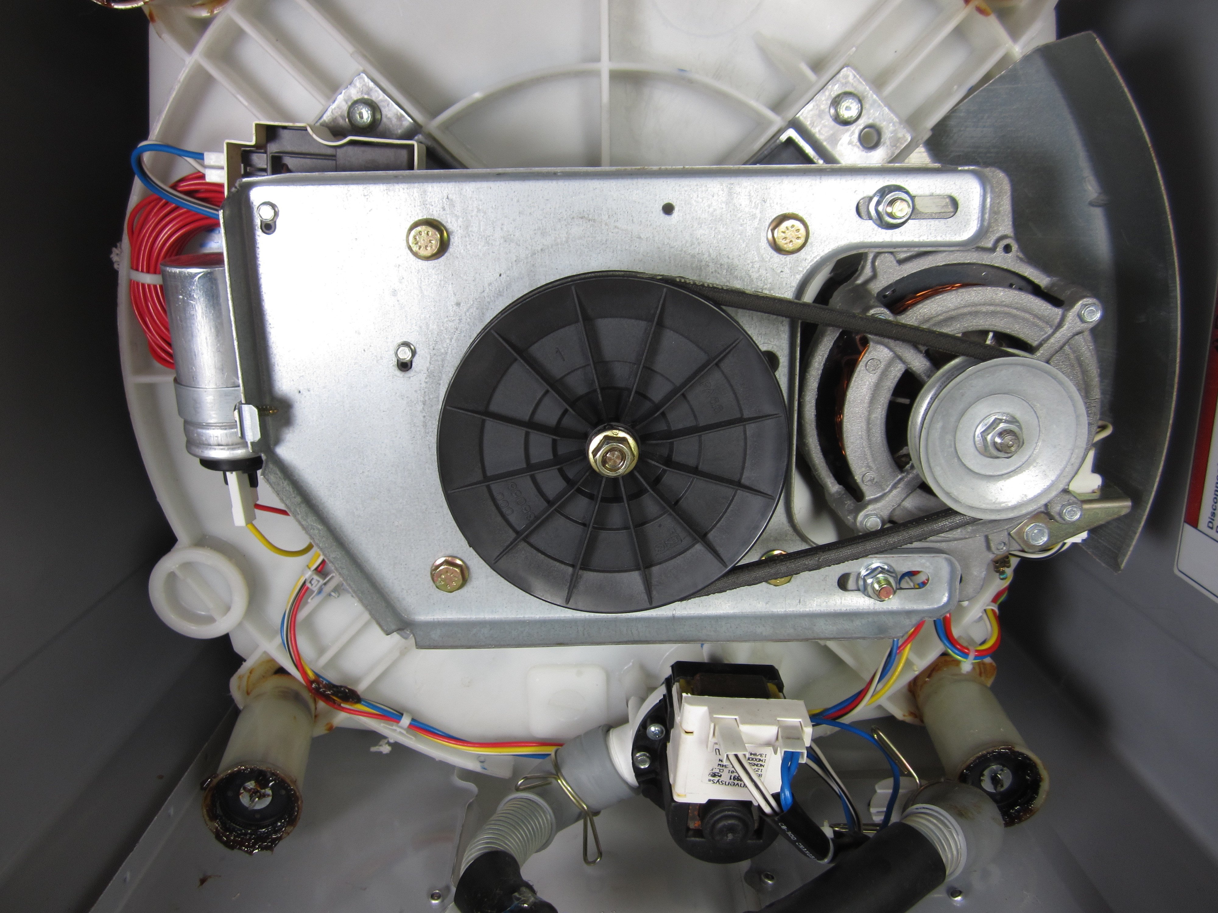 A real photo of the underside of a washing machine showing a black rubber drive belt looped around a large pulley and a smaller motor pulley