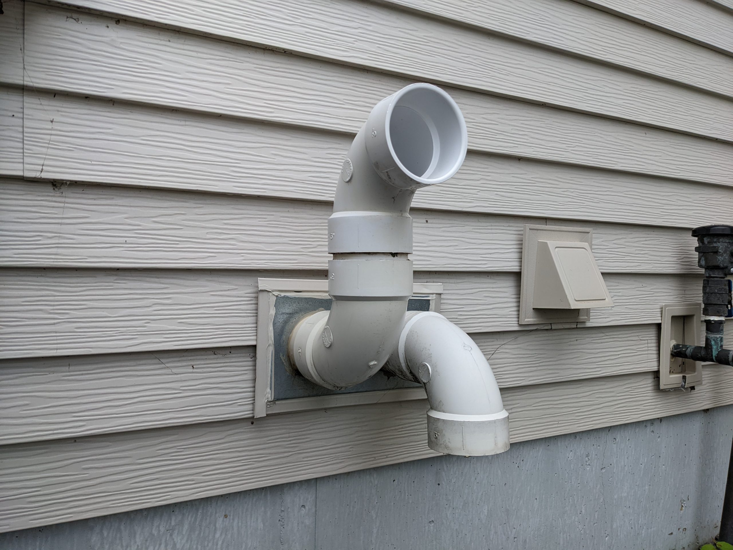 A real photo of two white PVC furnace pipes exiting the side of a house outdoors, showing the intake and exhaust terminations near ground level