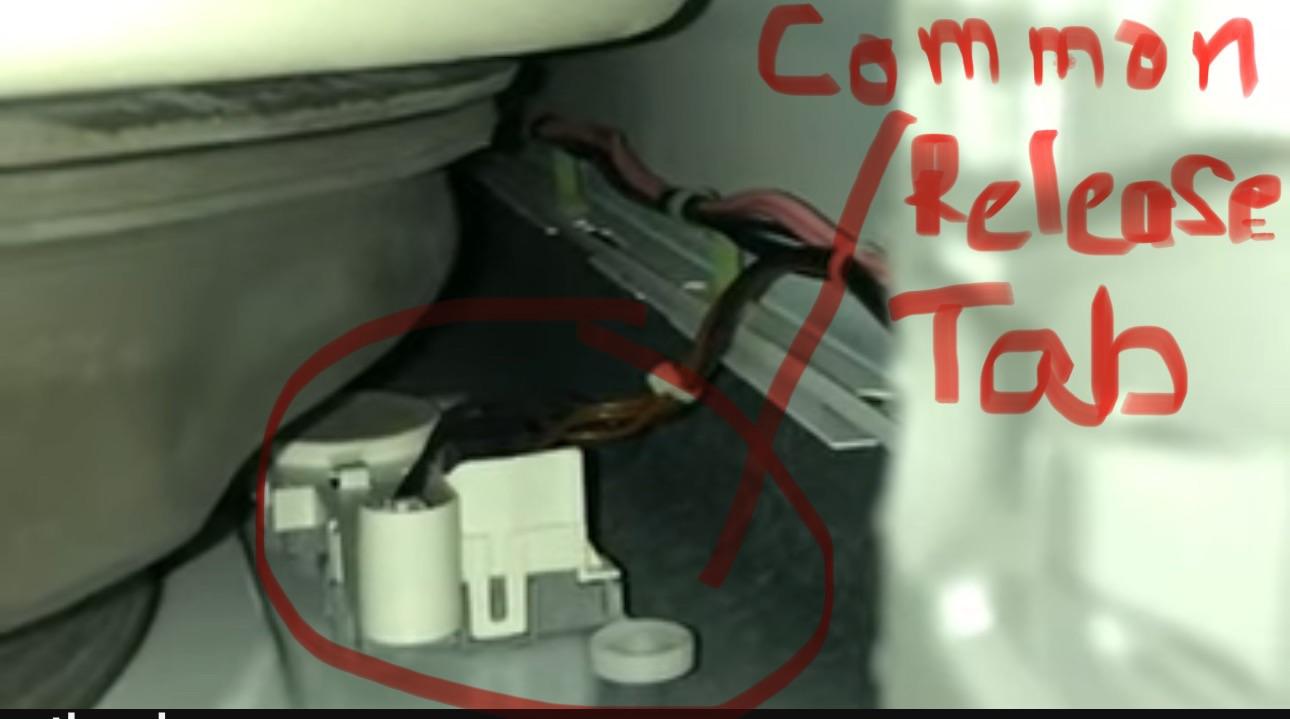 A real photo showing a hand reaching behind the lower access panel of a front-load washer to pull a small plastic emergency door release tab near the drain pump