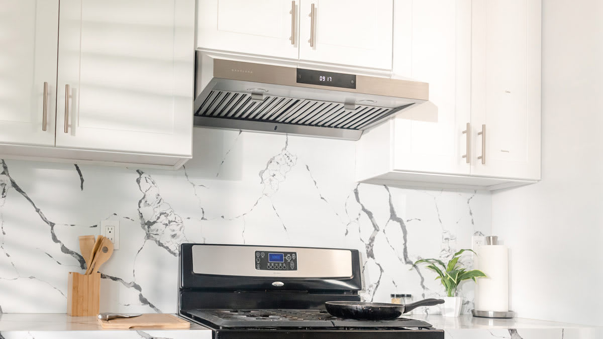 A real photograph of a home kitchen stove with the range hood lights on and steam being pulled upward while someone cooks