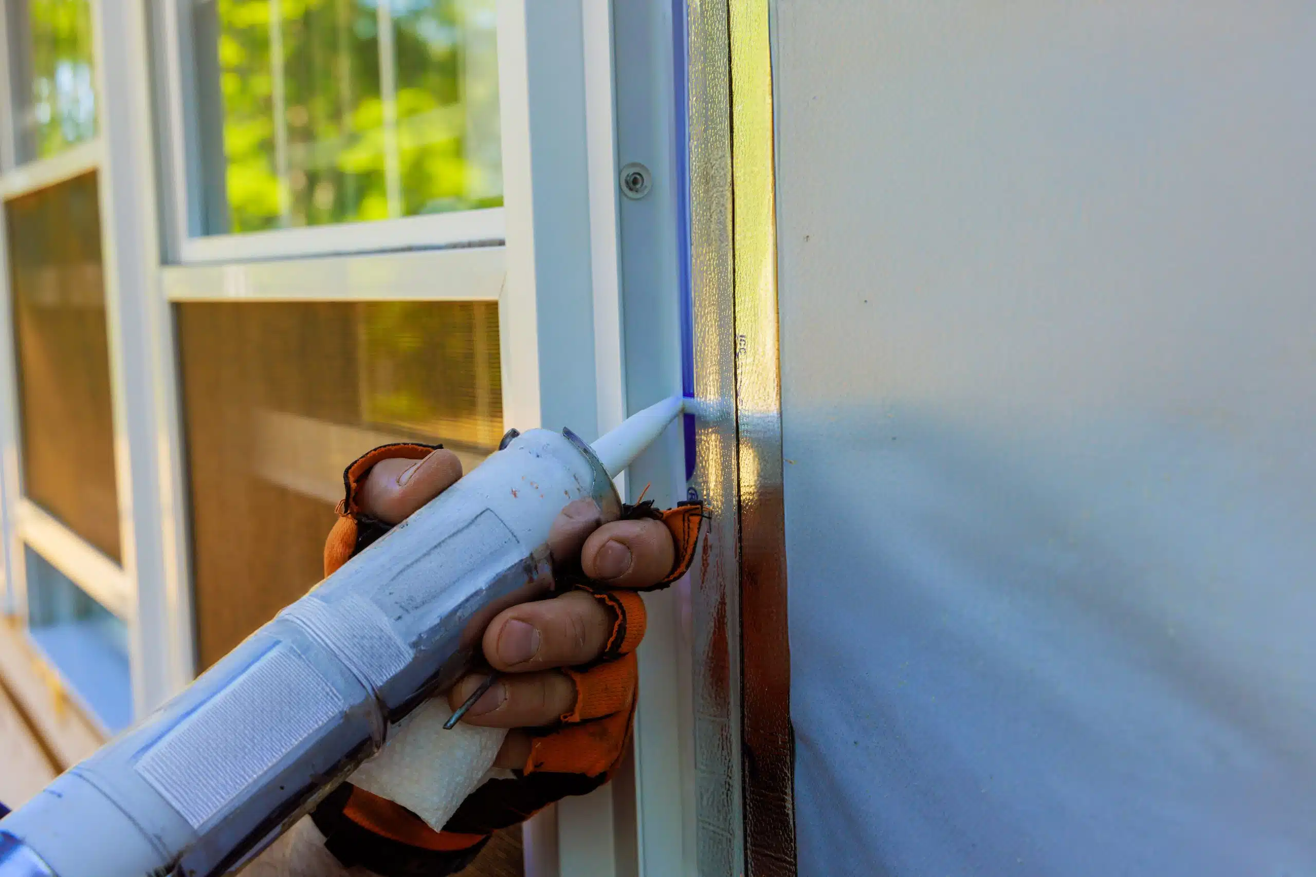 A real photograph of a homeowner’s gloved hands applying exterior-grade caulk along the edge of a window trim with a caulk gun on a house exterior in daylight