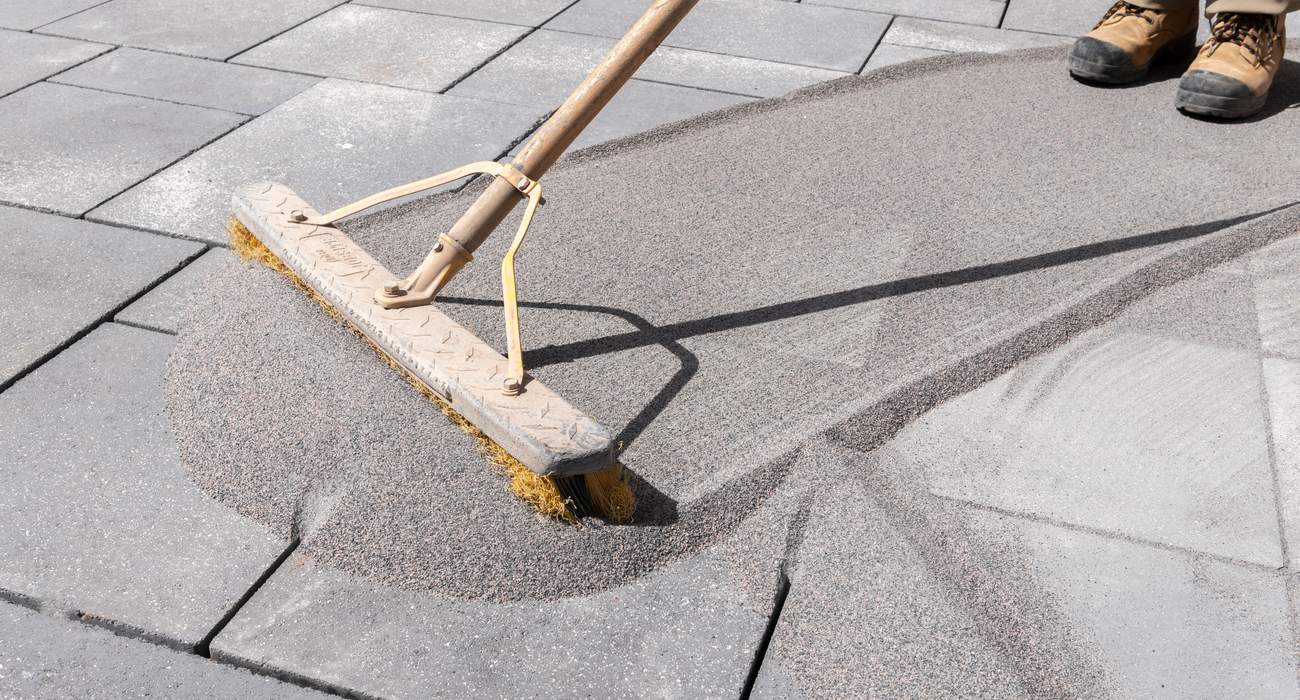 A real photograph of a person sweeping polymeric sand into paver joints on a patio, with a push broom angled across the pavers and sand filling the gaps