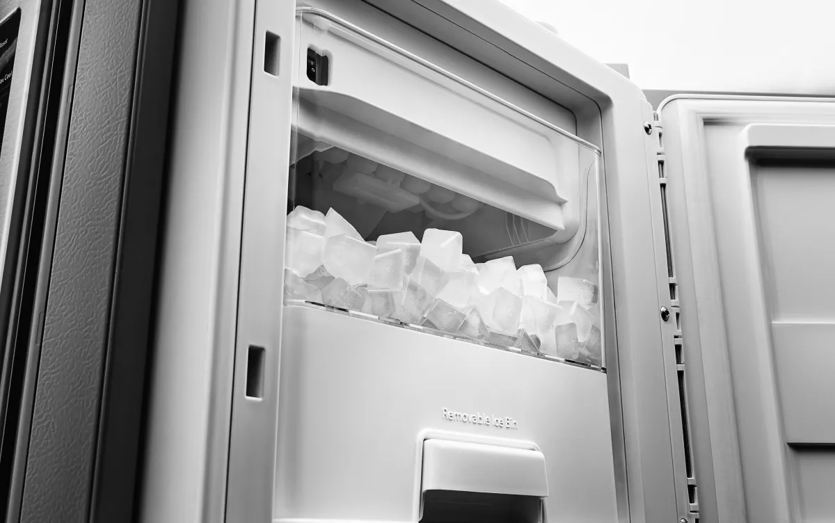 A real refrigerator freezer compartment with a built-in ice maker that is heavily frosted over, with ice buildup around the ice mold and surrounding plastic housing