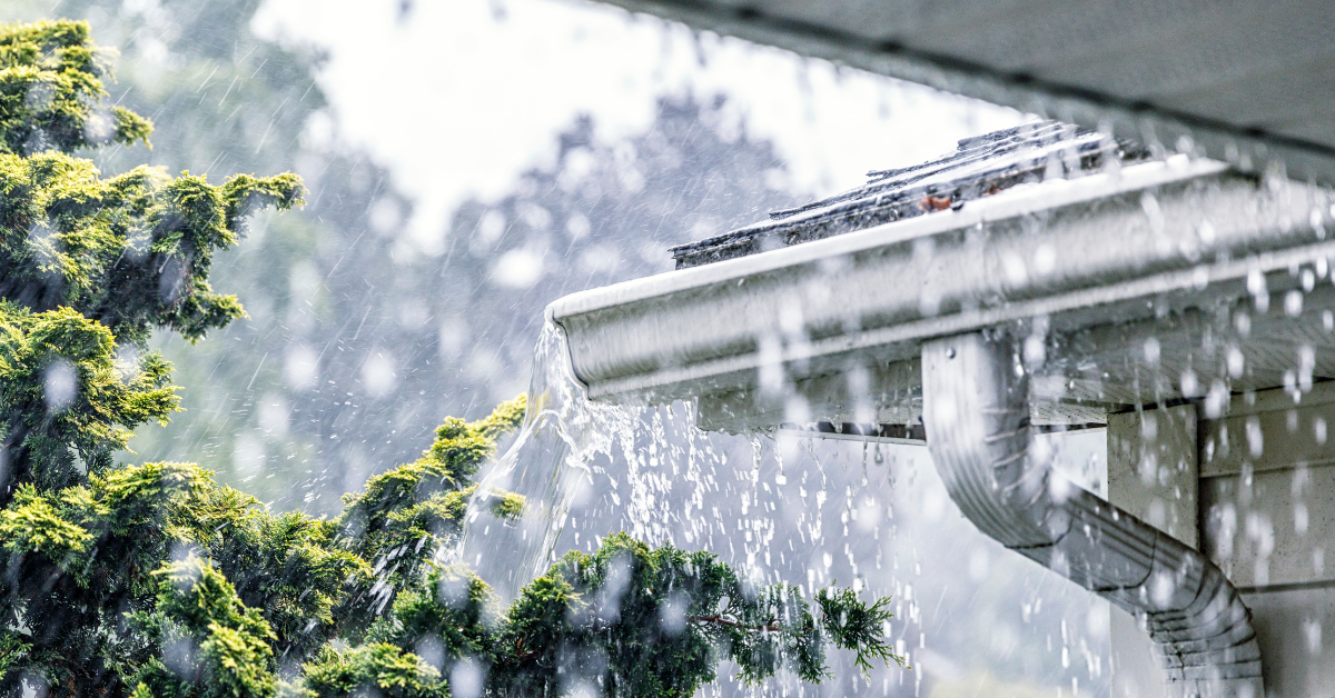 A real single-family home during a heavy rainstorm with water spilling over the edge of an aluminum gutter near a corner, splashing onto the ground below