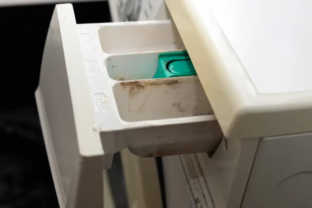 A real washing machine detergent drawer removed and held over a sink, showing dried detergent buildup in the compartments