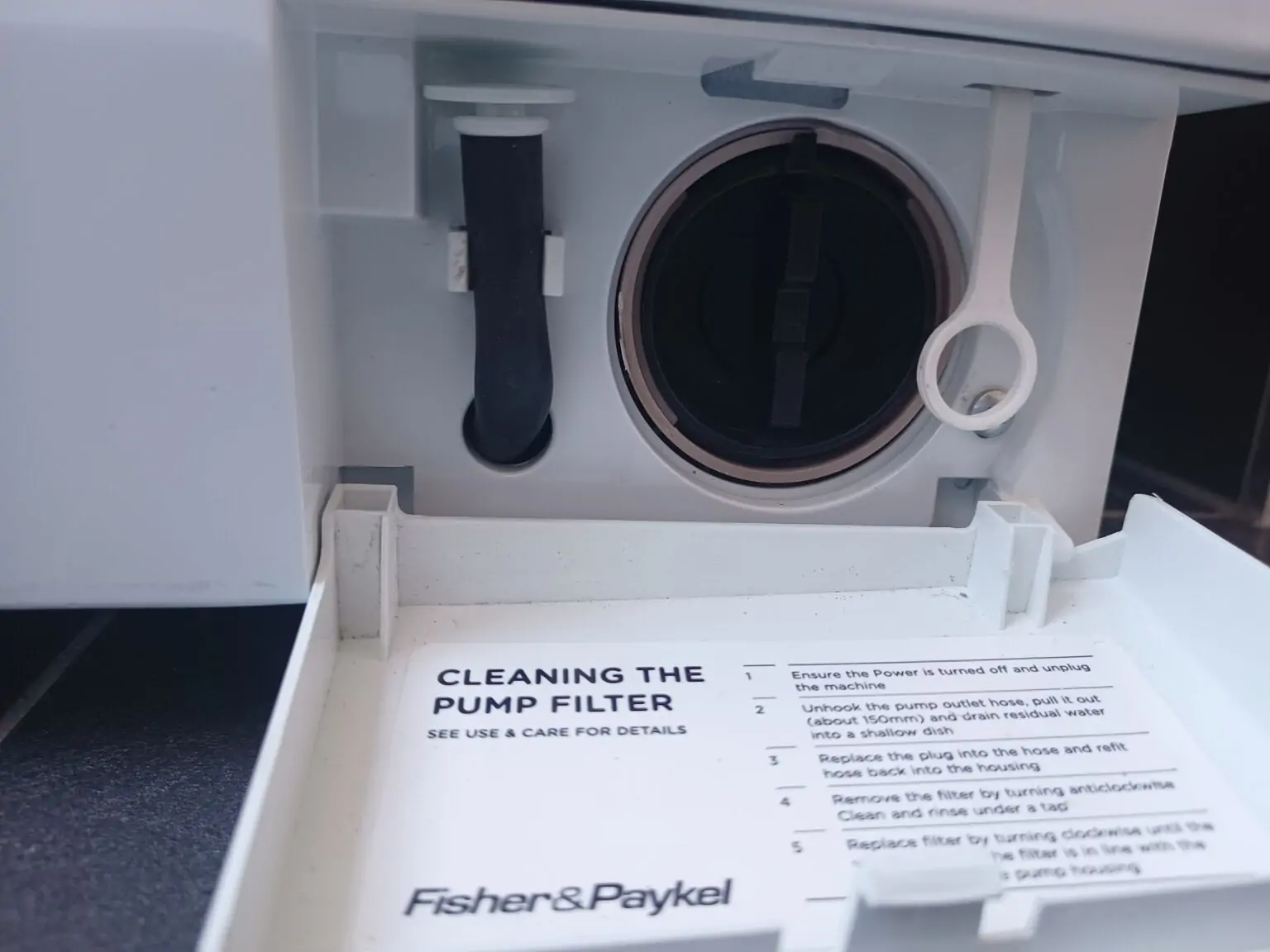 A real washing machine lower access door open with a person unscrewing the drain pump filter while towels and a shallow pan catch water