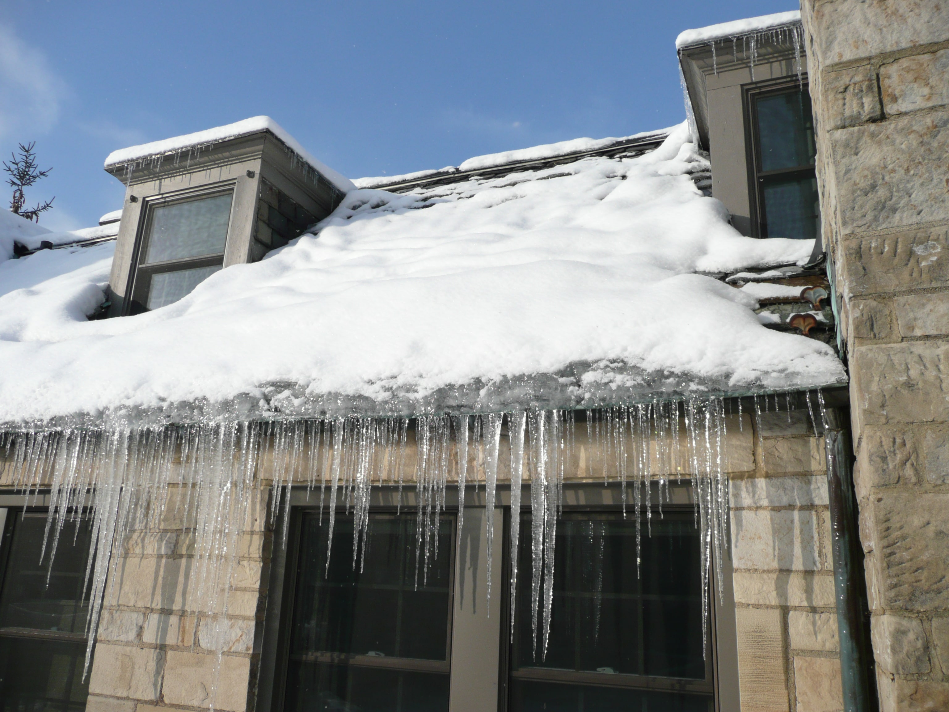 A real winter photo of a suburban house roof edge with a thick ridge of ice at the eaves and long icicles hanging over the gutter