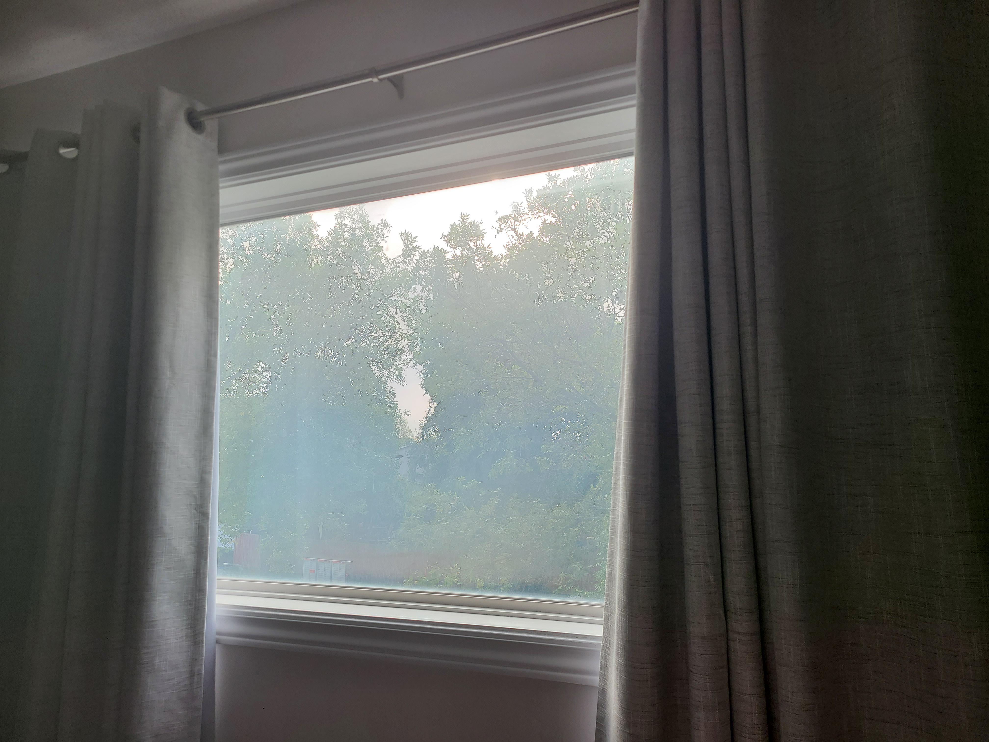 A realistic photo of a modern double-pane window with a cloudy, hazy patch trapped between the panes that cannot be wiped away, viewed from inside a home