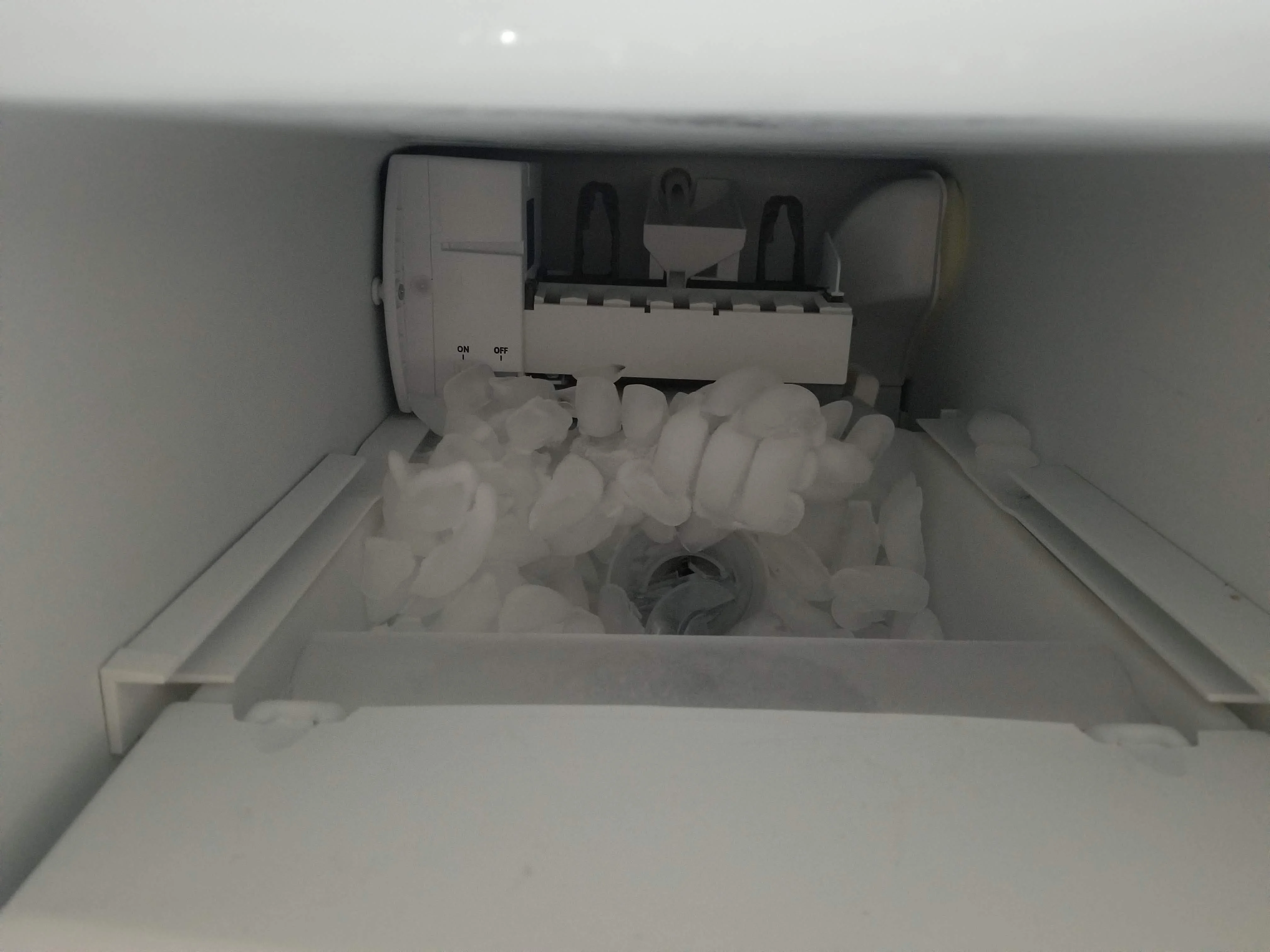 A refrigerator ice bin sitting in a kitchen sink with a solid clump of ice visible inside the bin, real photo