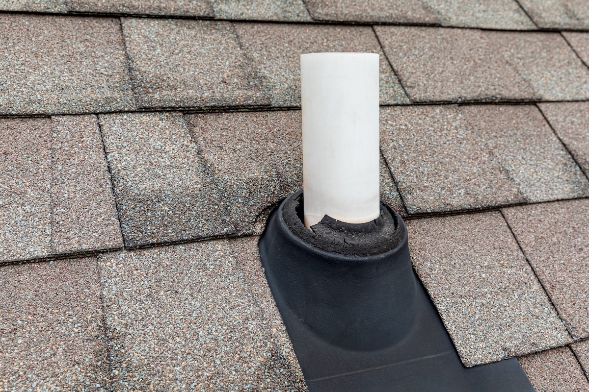 A residential asphalt-shingle roof with a single white PVC plumbing vent pipe sticking up near the ridge on a clear day, real photo