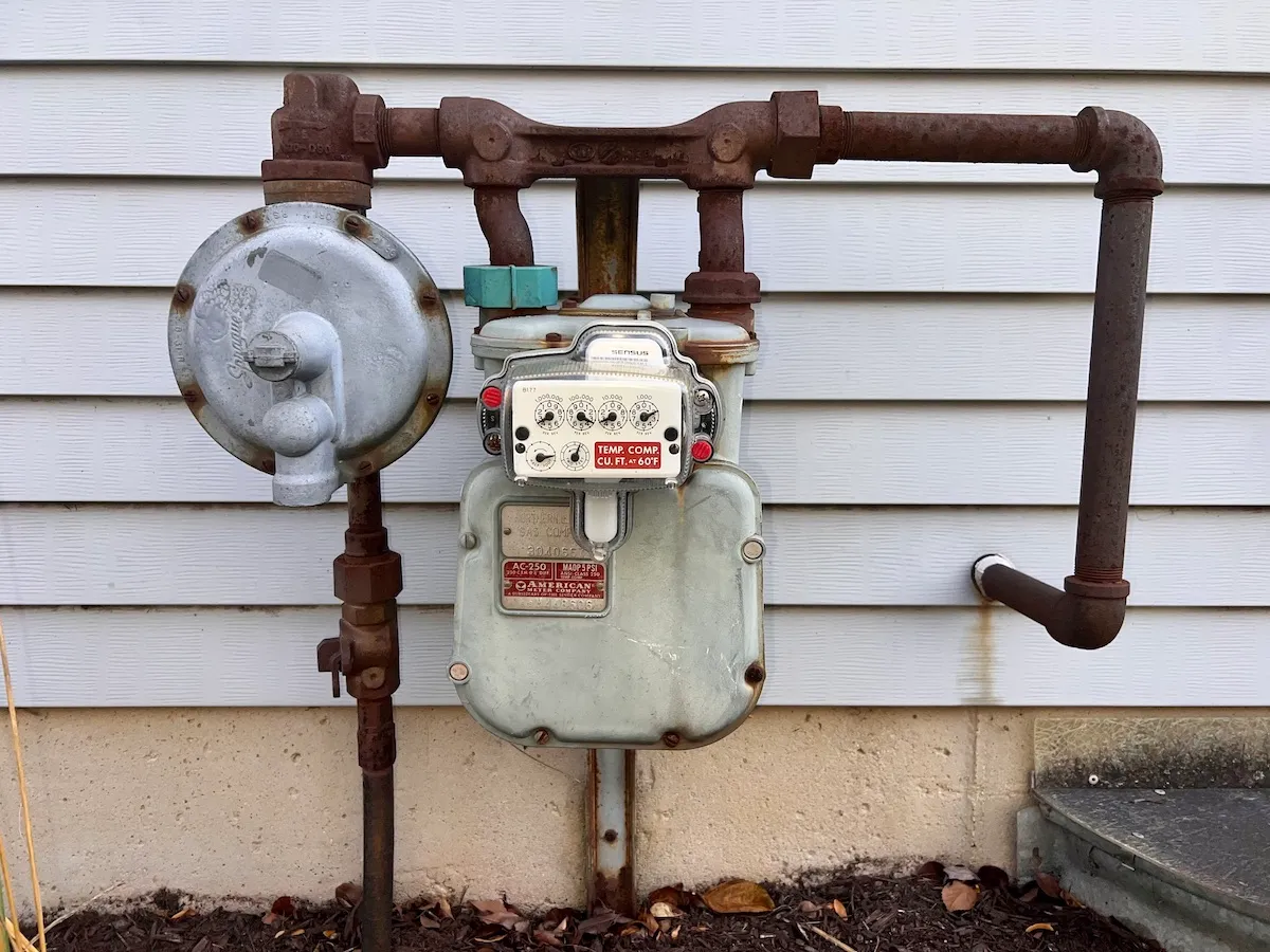 A residential natural gas meter mounted on an exterior wall with a visible shutoff valve and pipe connection, real photo style