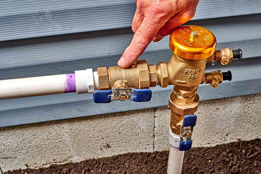 A residential pressure vacuum breaker backflow preventer installed on a side yard irrigation line with sunlight and a house wall in the background