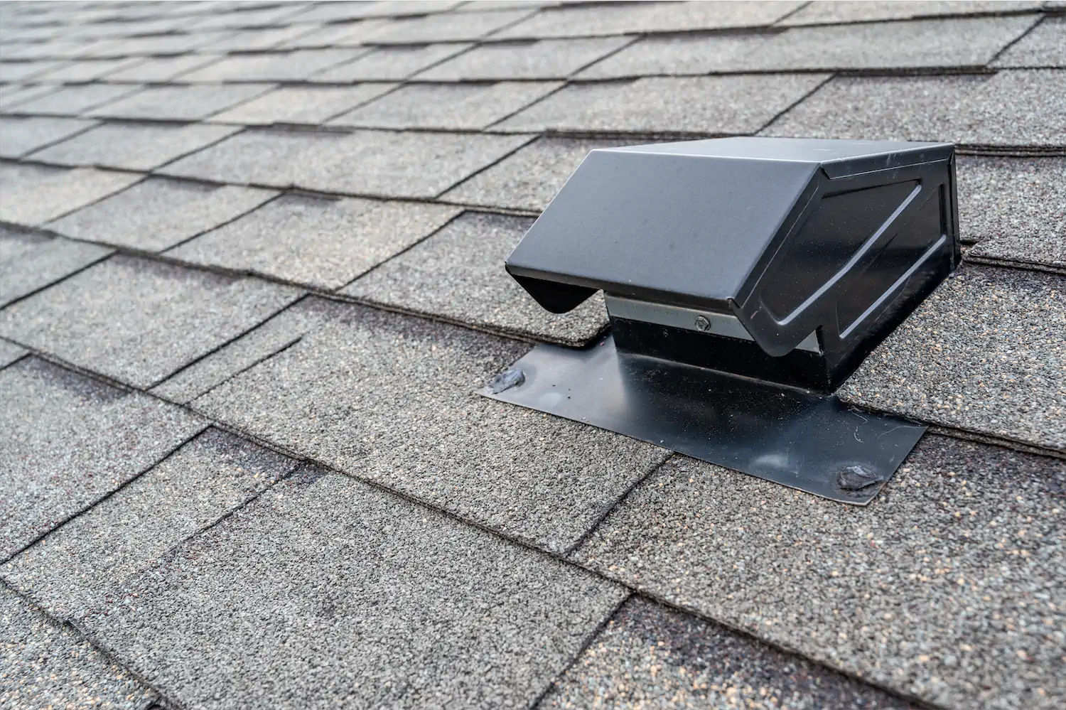 A roof-mounted attic exhaust fan housing sitting on an asphalt shingle roof on a sunny day, real photo