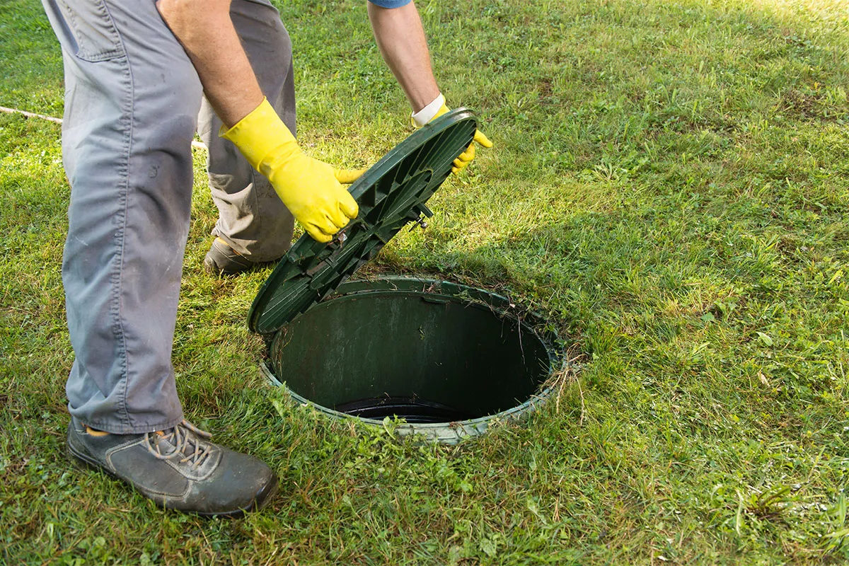 A septic service technician in work clothes kneeling in a grassy yard and inspecting an exposed septic tank access lid near a suburban home, real photo