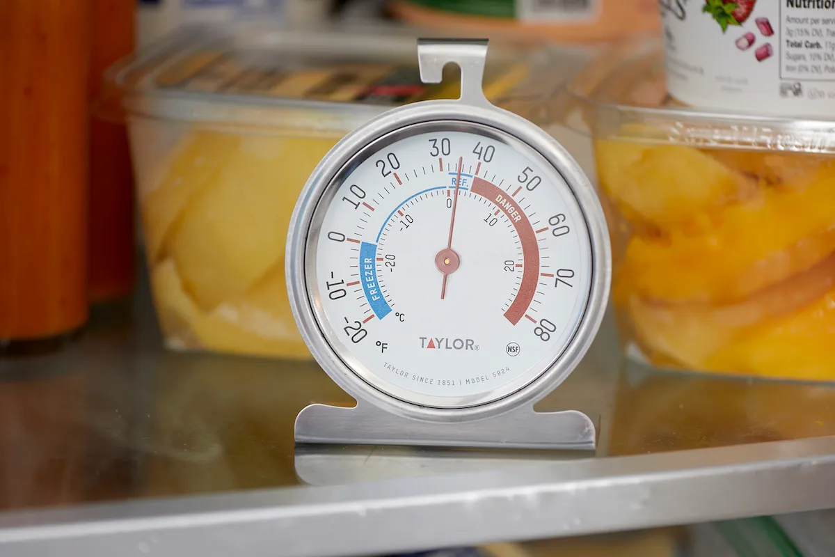 A simple dial freezer thermometer resting on a basket inside an open chest freezer, showing temperature measurement in progress