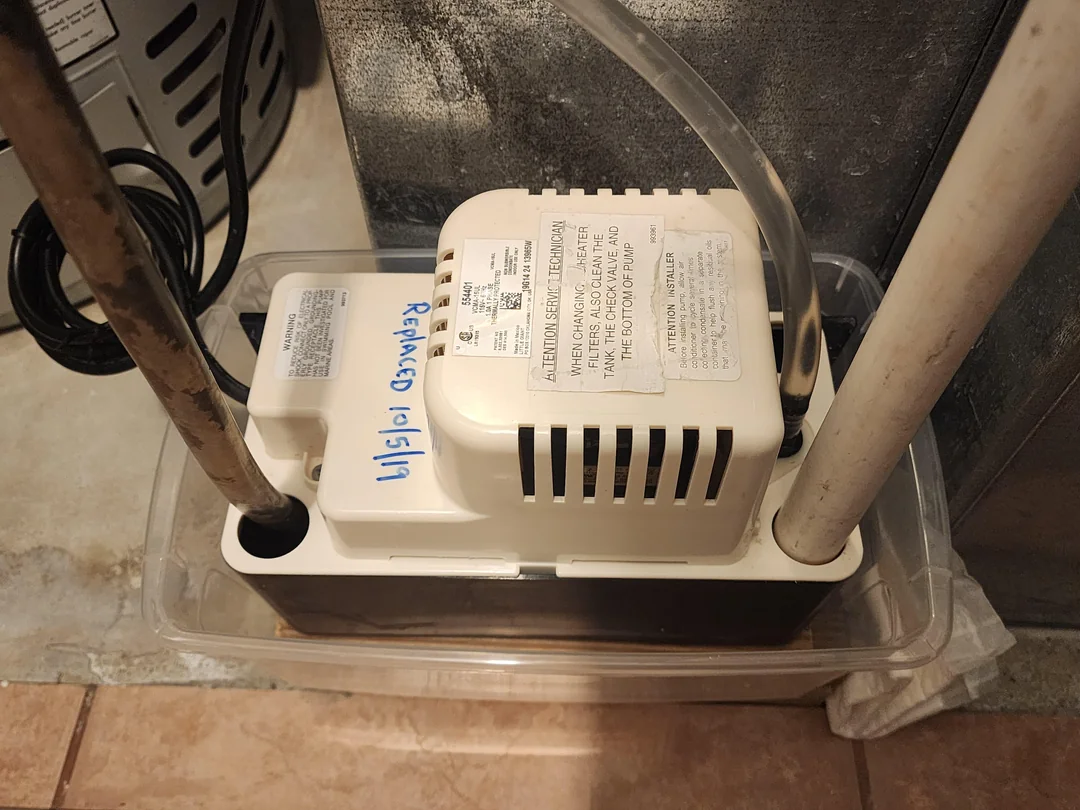A small condensate pump with a clear reservoir sitting on the floor next to a gas furnace in a utility room, realistic home photo