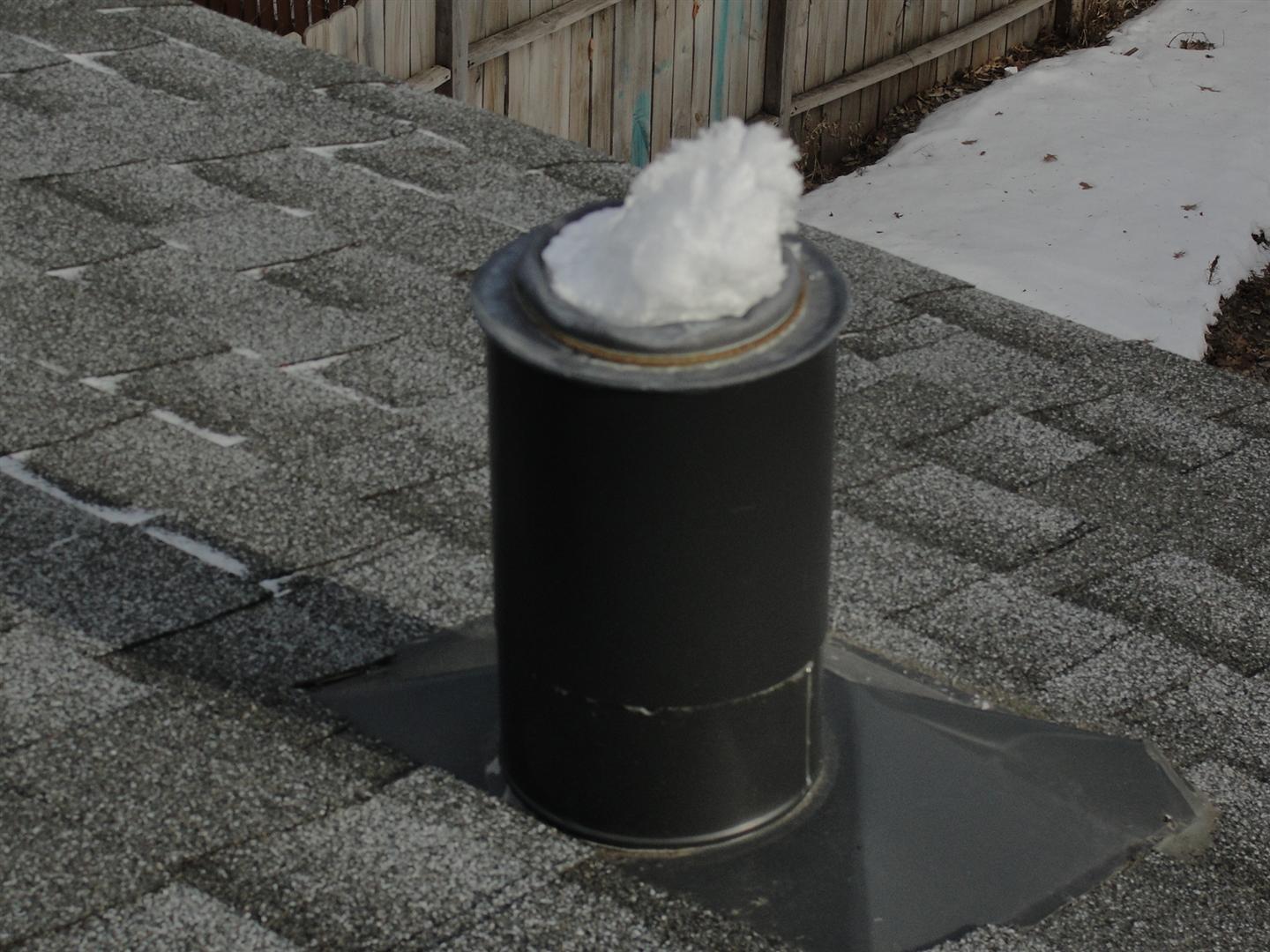 A snowy residential roof with a plumbing vent pipe showing a visible ice buildup around the opening, real photo
