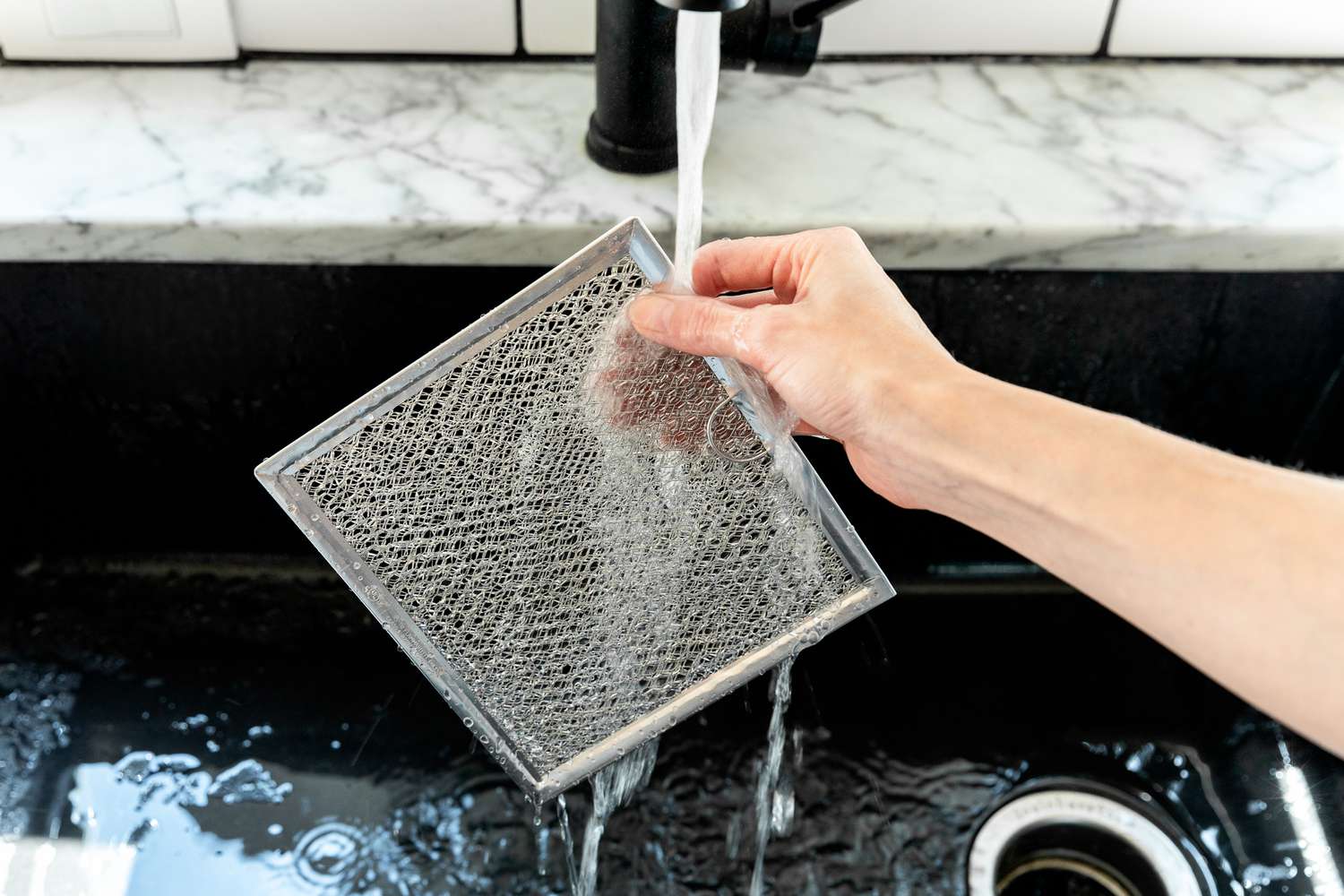 A stainless kitchen sink filled with hot soapy water soaking two metal microwave grease filters