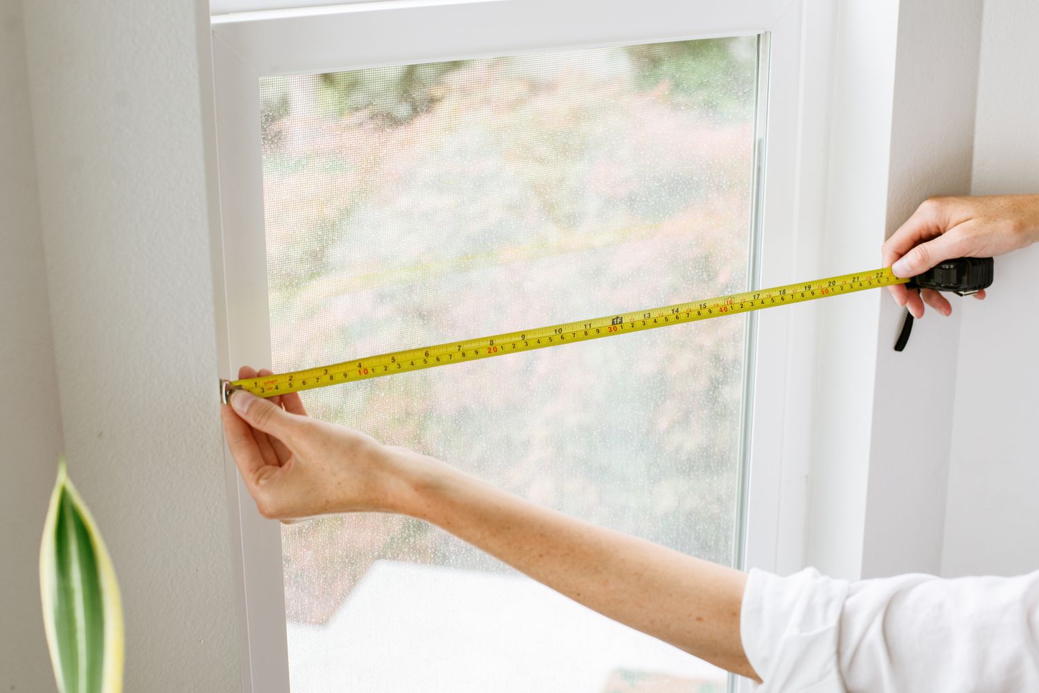 A tape measure stretched across the inside of a window frame at mid-height, showing how to measure width for an inside-mounted cellular shade