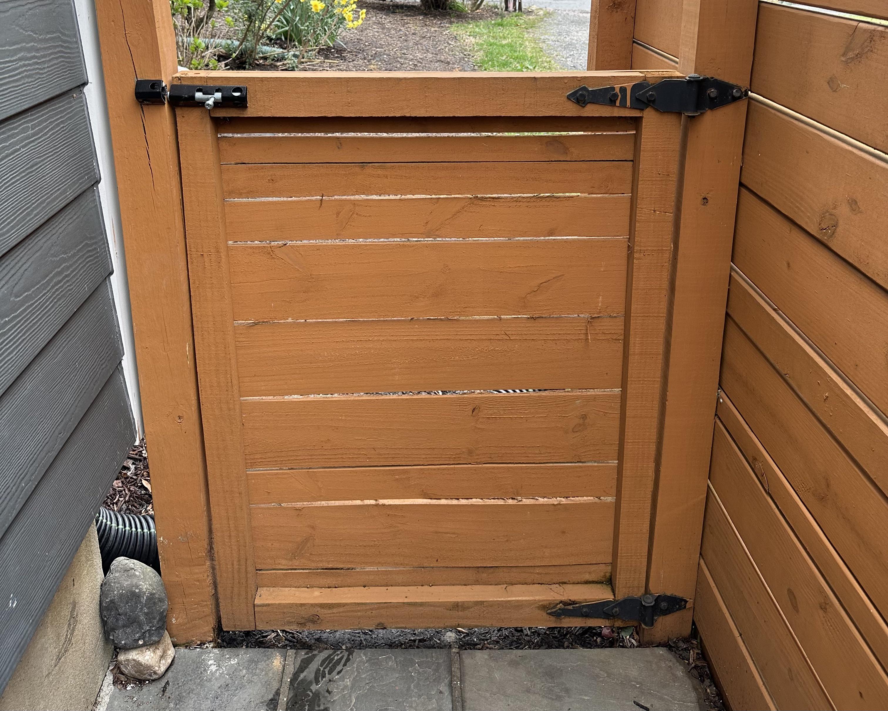 A wooden fence gate slightly sagging with visible hinges on the post, latch side dragging near the ground, backyard setting, photorealistic