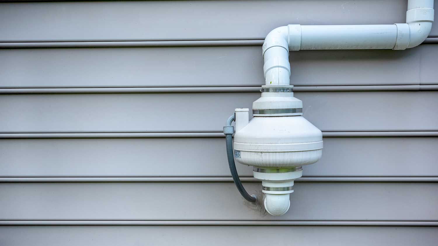 An exterior photo of a radon mitigation PVC vent pipe rising along a house wall with an inline radon fan mounted above, clear daylight
