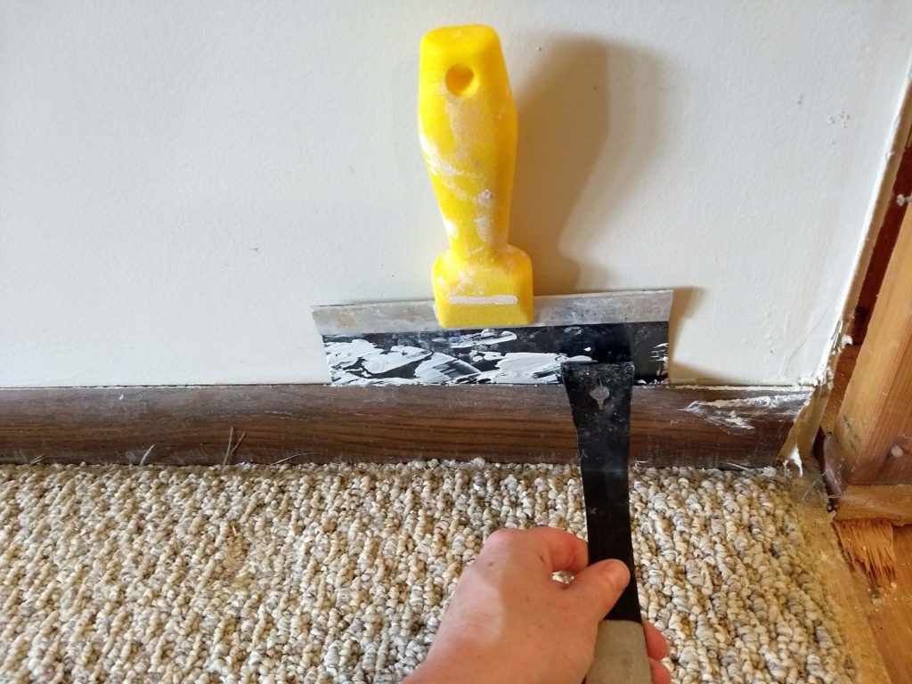 Close-up of a putty knife protecting a painted drywall surface while a small pry bar gently lifts an old baseboard, with the tool leverage pressed against the putty knife instead of the wall