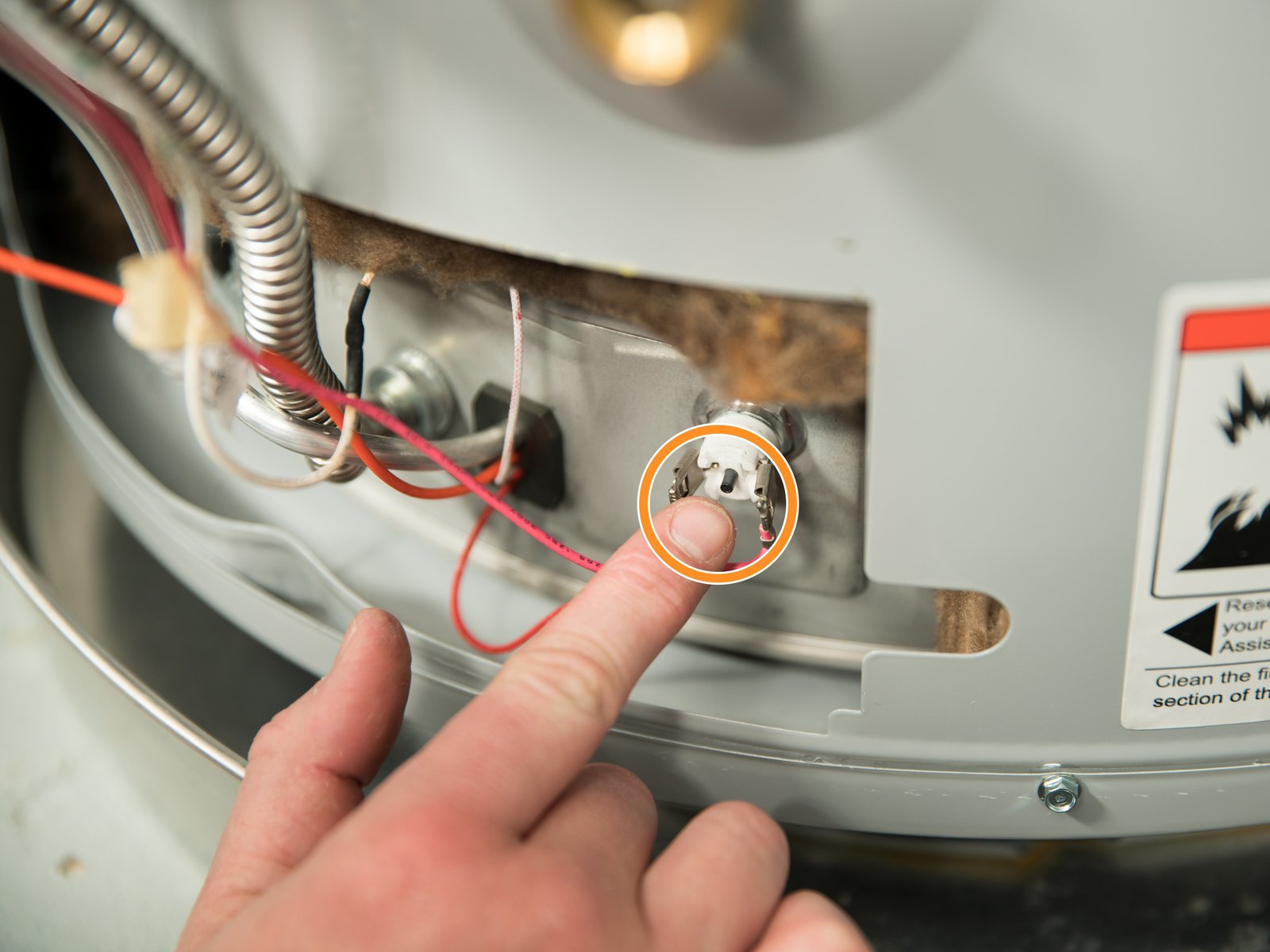 Close-up photo of an electric water heater upper thermostat behind an access panel with the small red reset button visible, natural workshop lighting