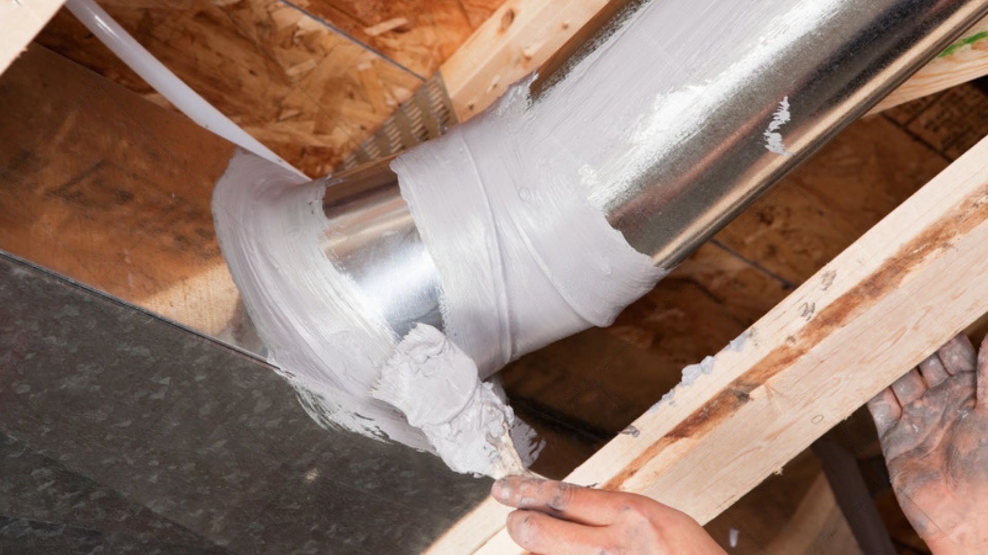 Hands applying HVAC duct mastic with a brush to seal a joint on metal ductwork in an attic