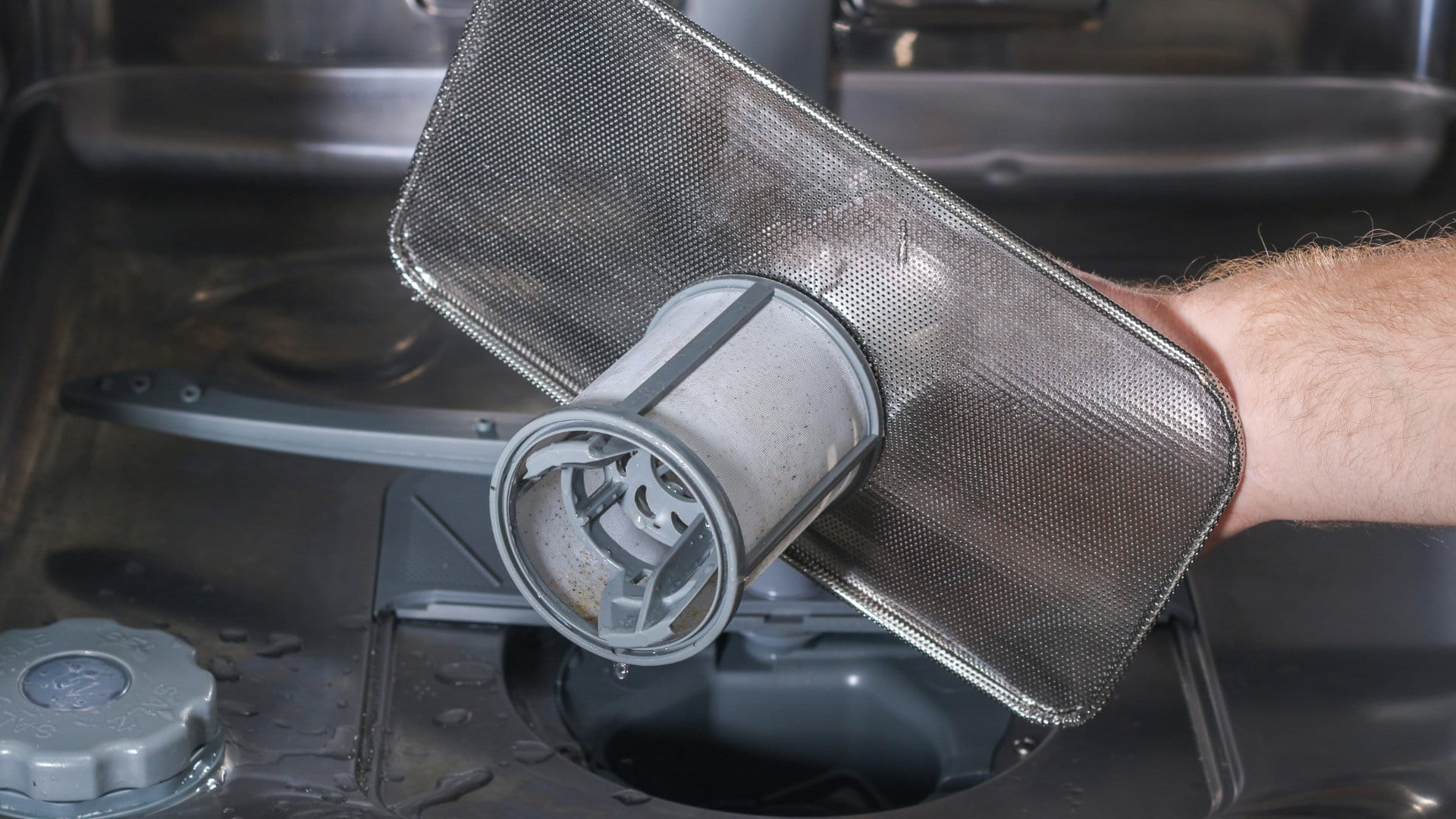 Hands lifting a cylindrical dishwasher filter out of the bottom of a stainless dishwasher tub