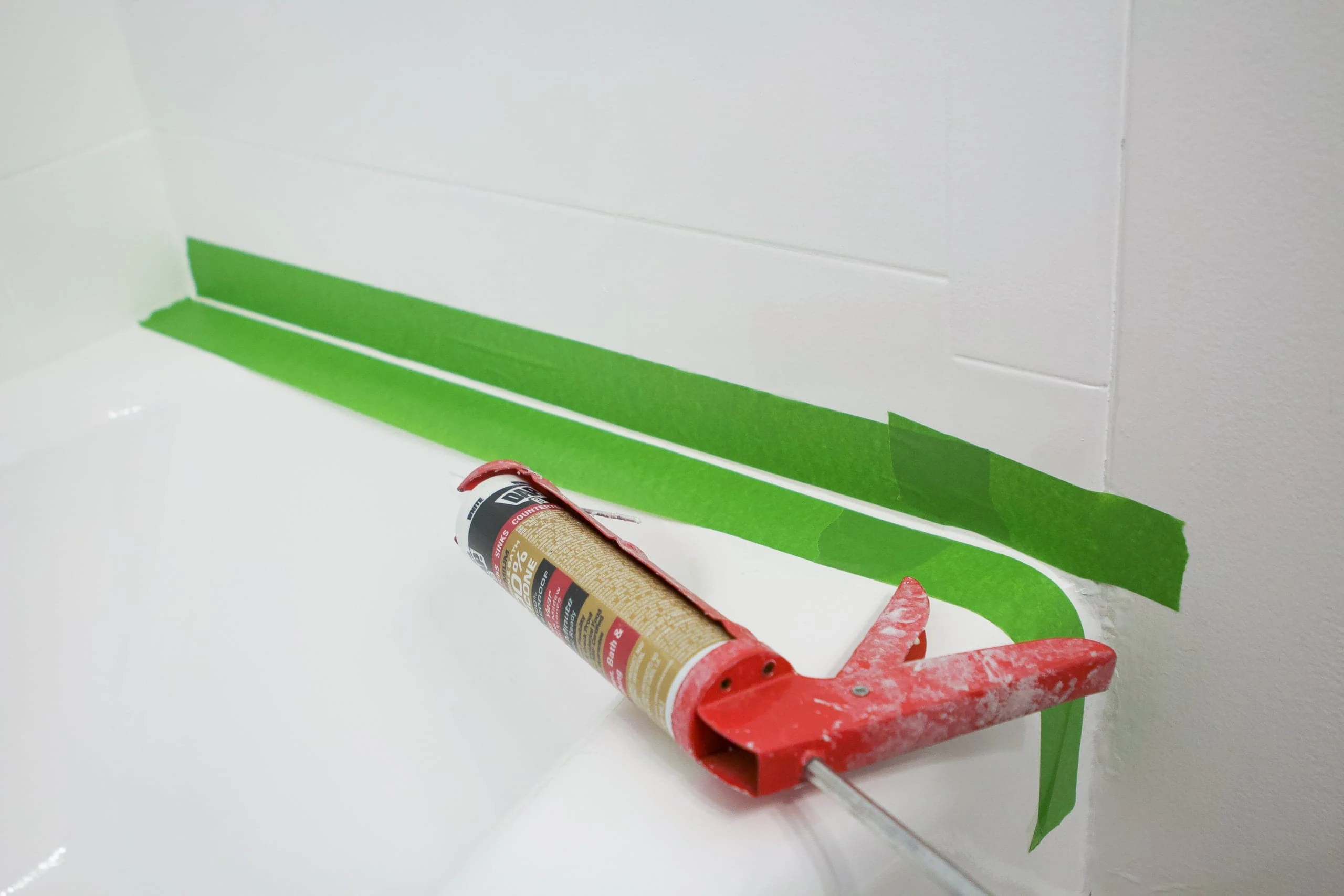 Painter's tape applied in two straight lines along a bathtub-to-tile seam leaving a narrow gap for caulk, close-up photo, photorealistic
